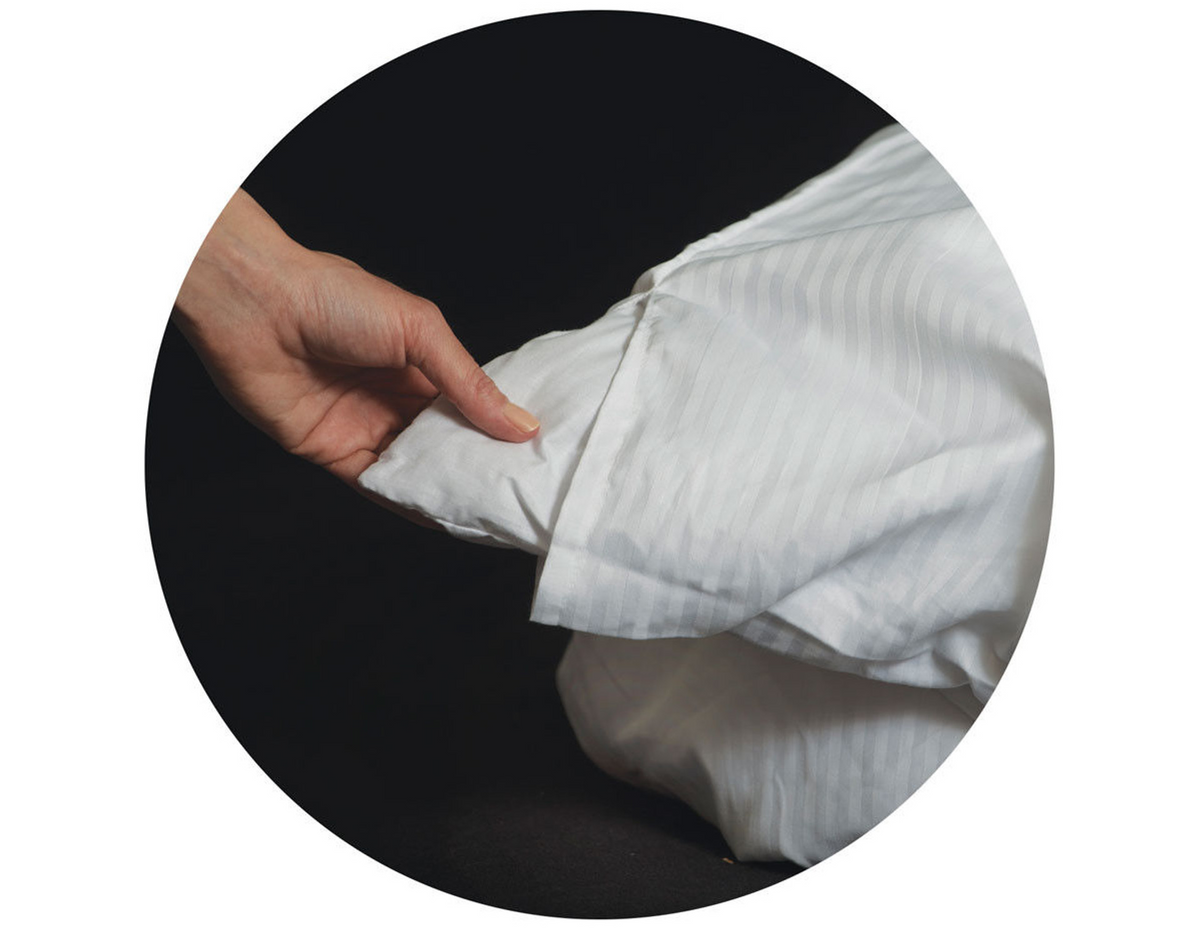 A hand tucks a Thomaston Mills Pillow Sham—310 Thread Count Satin Stripe—around a pillow against a dark background, shown inside a circular frame.