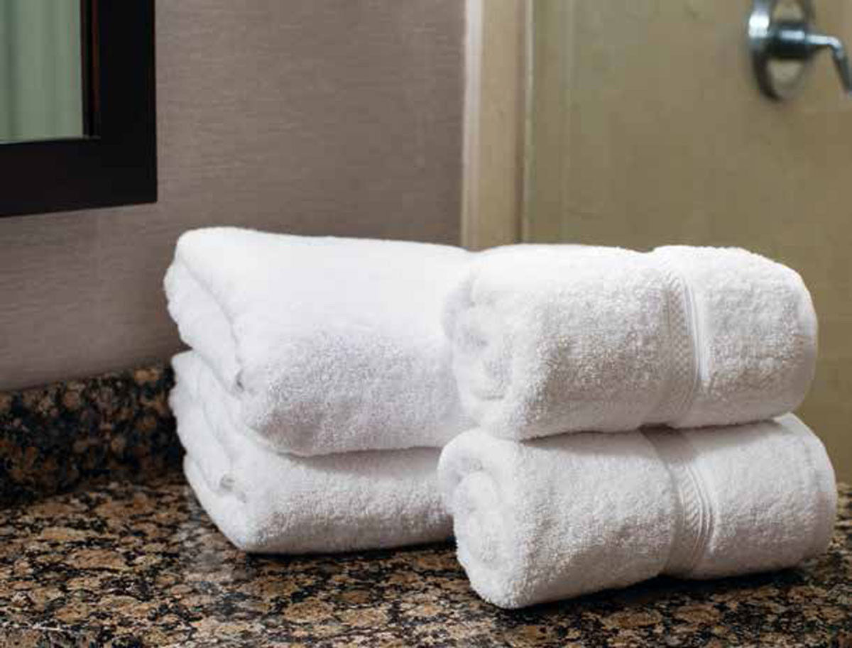 Two neatly folded and two rolled white Thomaston Mills Hand Towels, made from 100% ring spun combed cotton, rest on a dark granite bathroom countertop near a beige wall and a door.