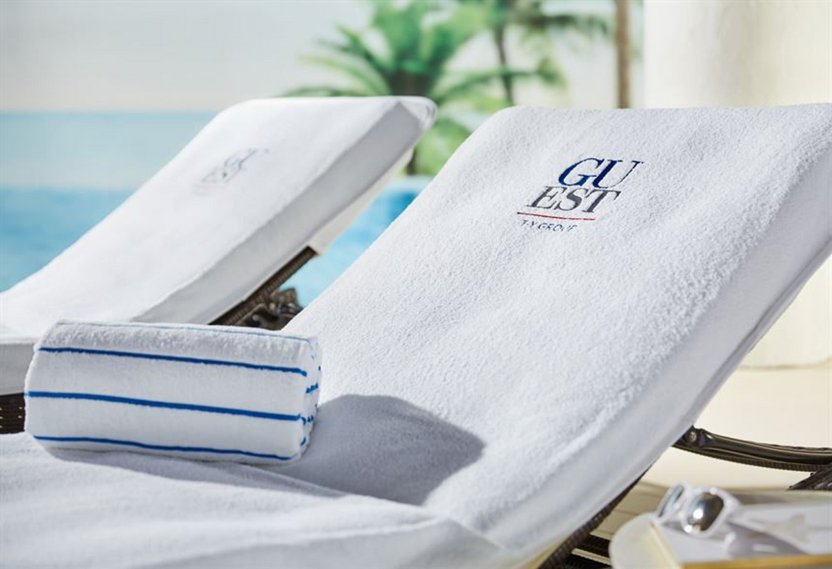 Two white lounge chairs by the pool feature 1Concier Loung Chair Covers and white towels embroidered with GUEST. A blue-striped rolled towel rests on one chair, with palm trees and water blurred in the background.