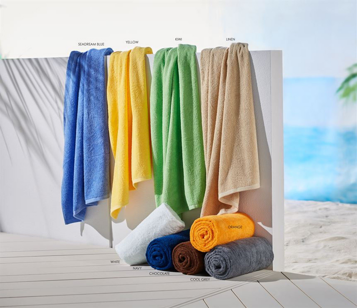 Six 1Concier Pool Towel Traditional 15 Lbs towels in blue, yellow, green, beige, white, navy, brown, orange, and grey are displayed on a white background with a blurred ocean and beach scene adding vibrant color.