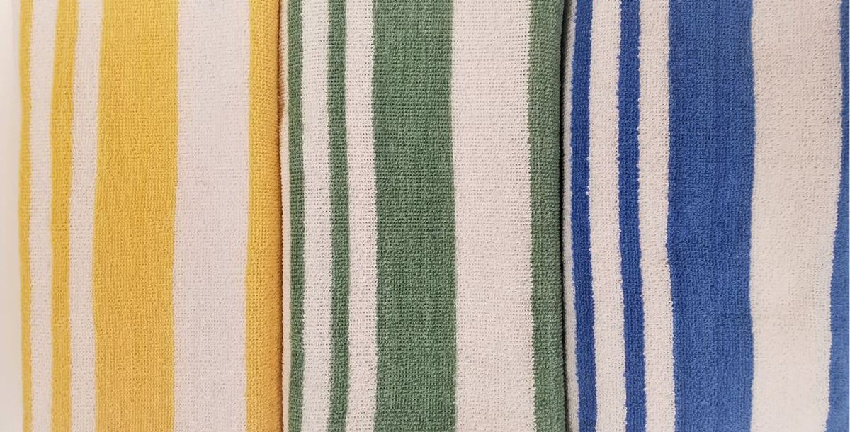 The Ganesh Mills Tropical Stripe Pool Towels, featuring vertical yellow/white, green/white, and blue/white patterns, are laid side by side—perfect cabana towels for relaxing by the pool.