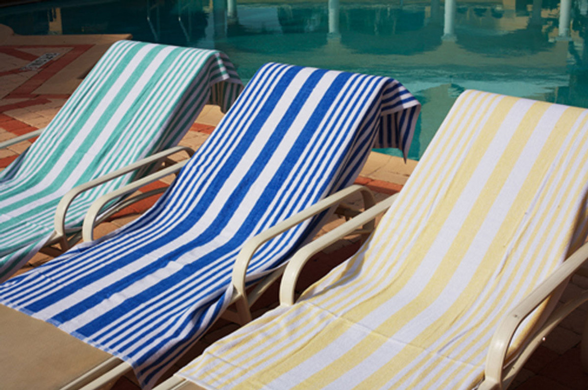 Three lounge chairs with Ganesh Mills Tropical Stripe Pool Towels in green, blue, and yellow are lined up on a tiled deck beside a sparkling swimming pool.