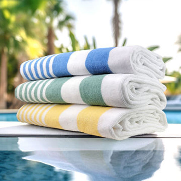 Three rolled Oxford Playa 2x2 Cabana Pool Towels by Ganesh Mills—striped in blue, green, and yellow—rest on a glass surface near a hotel pool, with blurred palm trees and lush greenery in the sunny background.