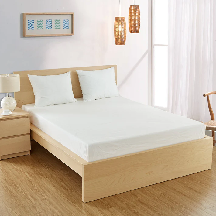 A neatly made bed with white bedding and two pillows features the Bargoose 6 Gauge Vinyl Fitted Waterproof Mattress Cover (9 deep). The cozy, sunlit bedroom has wooden floors, a nightstand with a lamp, wicker lights, and framed artwork.