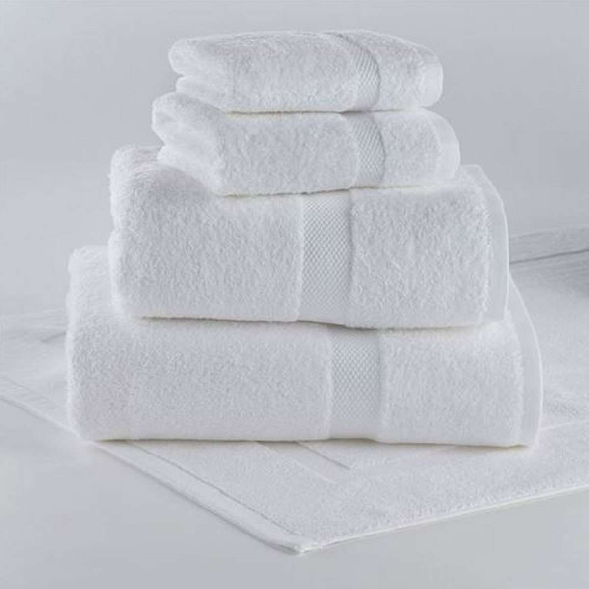 The Ruby Bath Towel by 1Concier, made from 100% cotton, sits plush and neatly stacked with washcloths on a matching white bath mat, all featuring an elegant rice weave border from the Ruby Collection, against a plain white background.