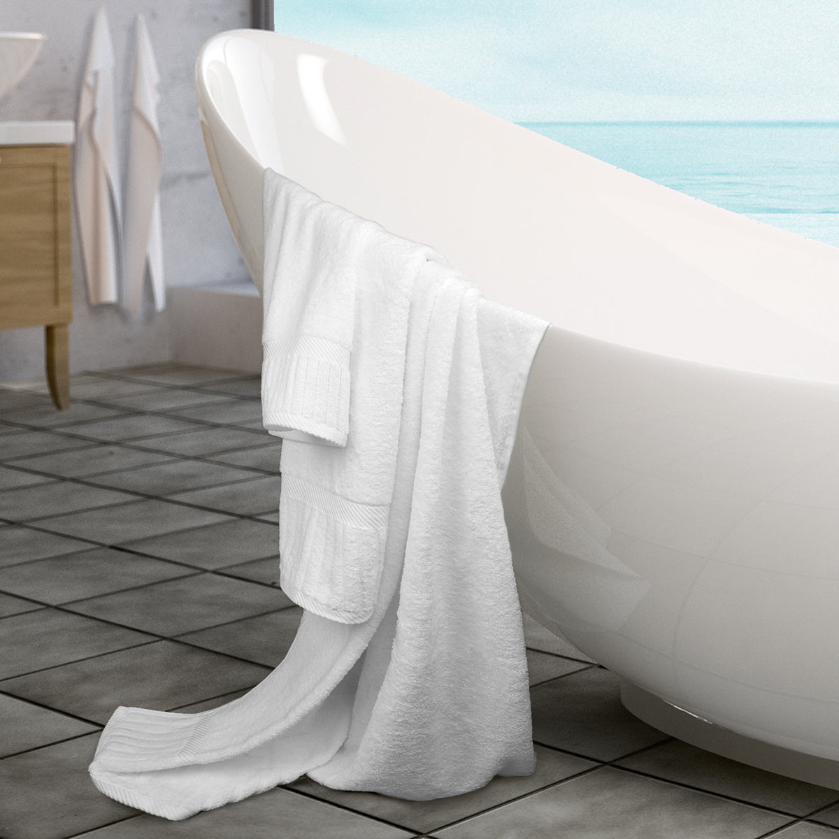 A large white bathtub sits on a tiled floor beside a window with a water view. A Ganesh Mills Washcloth - White Oxford Signature Towel and matching white towels hang over the tub’s edge, with another towel seen in the background.