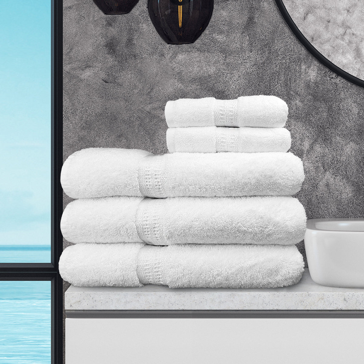 A luxurious Ganesh Mills Bath Mat - White Oxford Reserve Towel rests near a stack of matching towels on the bathroom counter by a white sink, with a window offering an ocean view beside the round mirror on the gray wall.