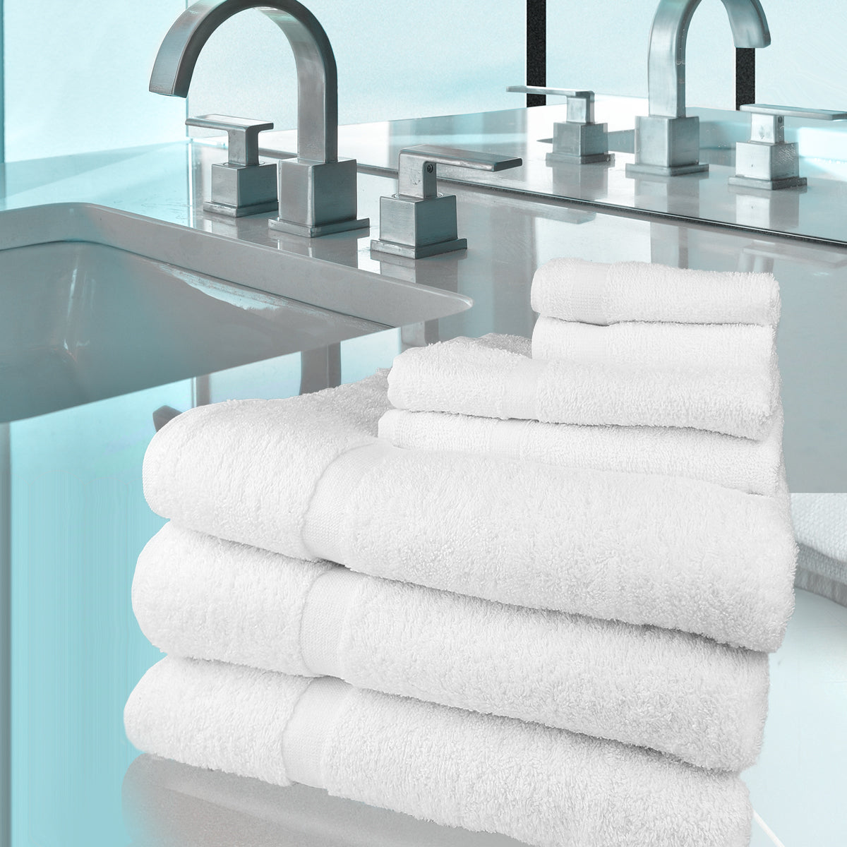 A stack of white Ganesh Mills Bath Towels from the Oxford Gold Cam Towel Collection sits on a bathroom counter near a modern sink and faucet, against a light blue background.