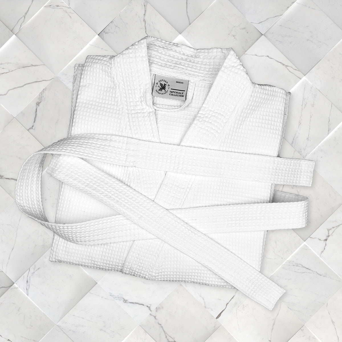 A neatly folded Ganesh Mills Oxford Honeycomb Waffle Bathrobe with a matching White Replacement Belt displayed on a white marble-tiled surface.