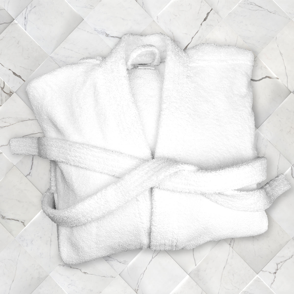 A Kimono - White Oxford Terry Bathrobe by Ganesh Mills, neatly folded with its belt, is displayed on a white marble tile background.