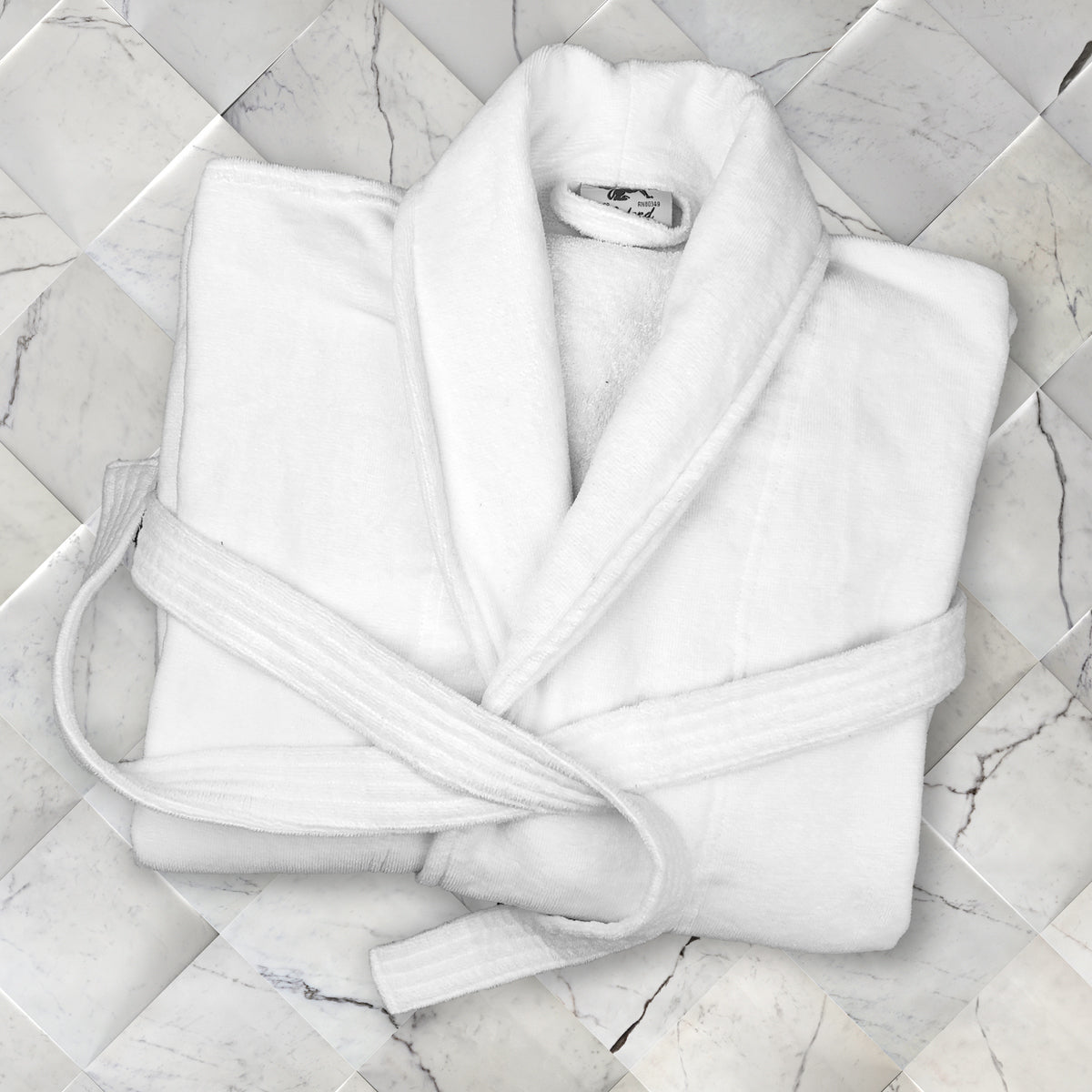 A Shawl Collar - White Oxford Velour Bathrobe by Ganesh Mills, neatly folded with its belt, rests on a light marble tile surface, exuding a clean and luxurious look.