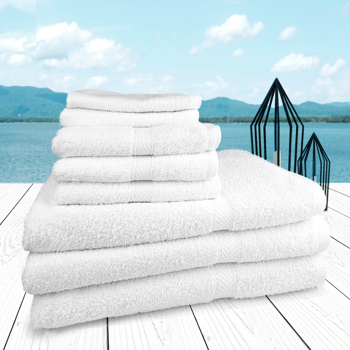 A neatly folded stack of Ganesh Mills Washcloth Square Hemmed - White Oxford Gold Dobby Towels rests on a white wooden surface outdoors, with a lake, mountains, blue sky, and black candle holders visible in the background.