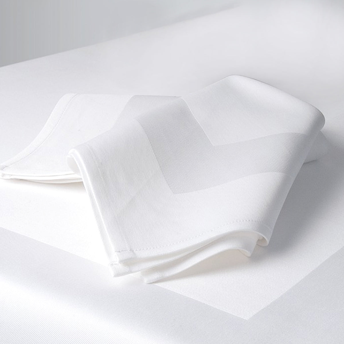 A neatly folded white napkin with a subtle zigzag pattern rests on a white tablecloth, highlighting the elegance of Ganesh Mills Napkins – White/Black Oxford Satin Band Damask Table Linens for a refined table setting.