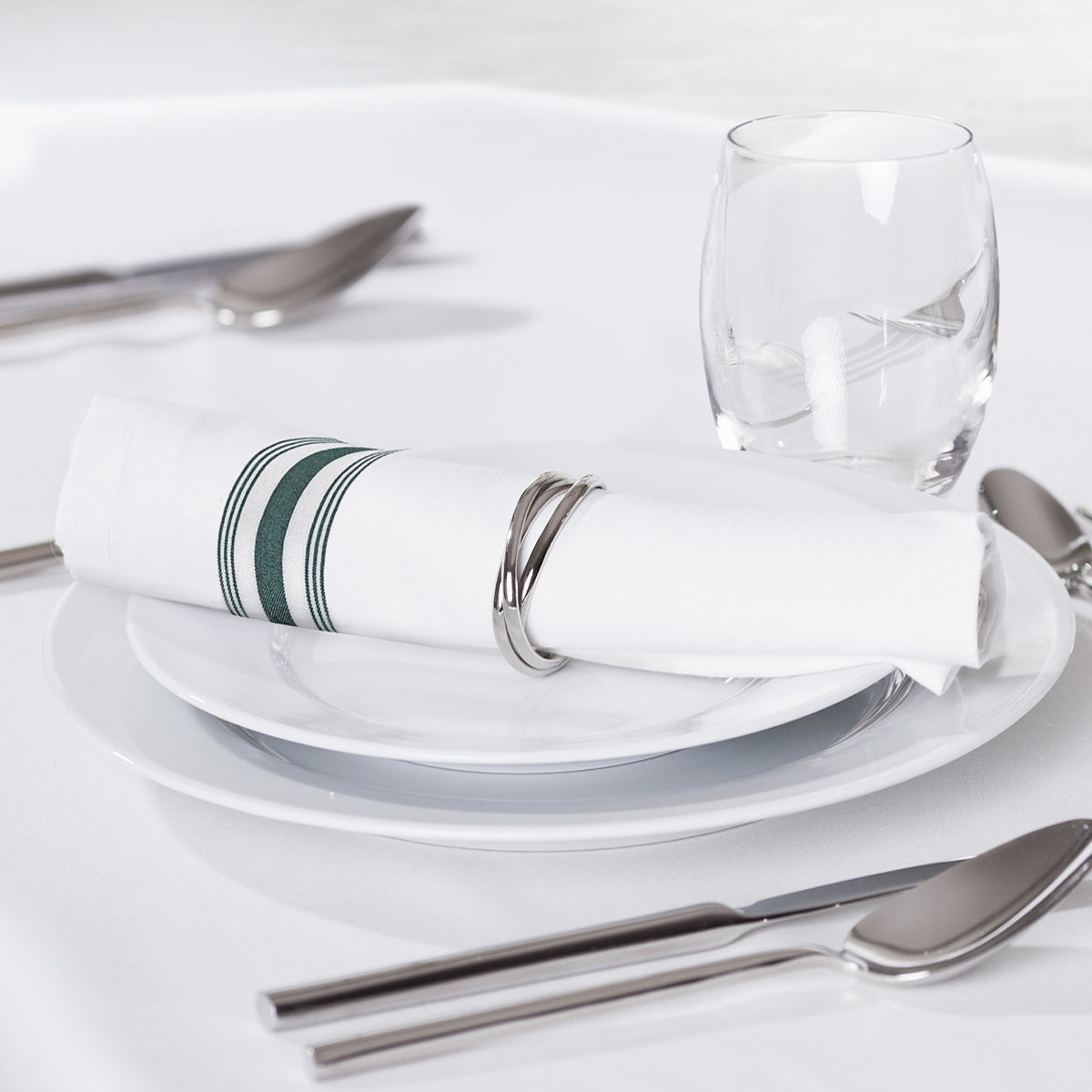 A neatly set dining table features a white plate, stainless steel cutlery, a glass, and a Ganesh Mills Bistro Napkin—white with color stripes—rolled in a silver napkin ring, all atop a white tablecloth.