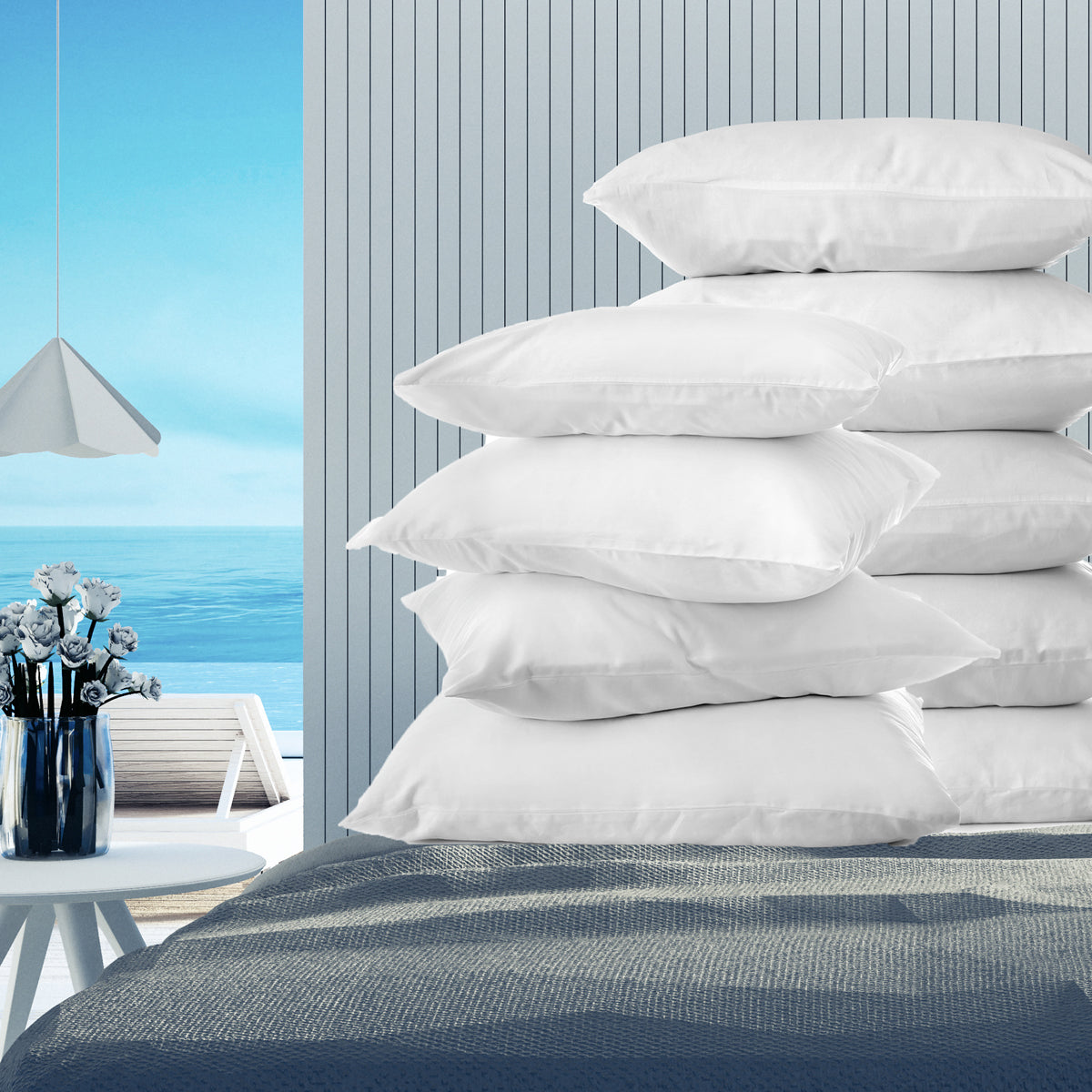 A stack of eight Ganesh Mills Pillow Cases with 3" Hem from the Oxford Super Deluxe Bed Linen collection sits on a bed in a modern, coastal room with blue walls, white flowers, and an ocean view, creating an inviting display.