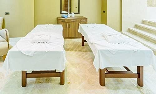Two massage tables with KSE Flat Sheets - Premium T130 (Twin/Twin XL) sit side by side in a spa room with soft, neutral tones. In the background, a wooden cabinet holds towels and bottles, and steps rise along the right wall.