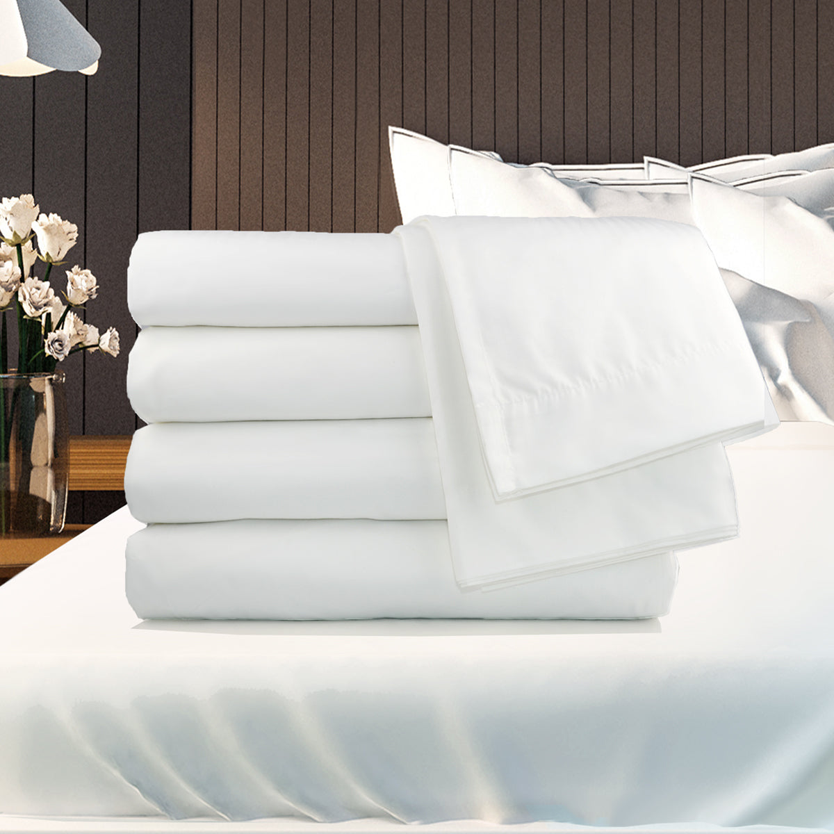 A neatly folded stack of Ganesh Mills Flat Sheets 2 Top Hem & 1 Bottom Hem - Oxford T180 Superblend Bed Linen is placed on a bed with white pillows in a modern bedroom, with a lamp and flowers on the wooden bedside table.