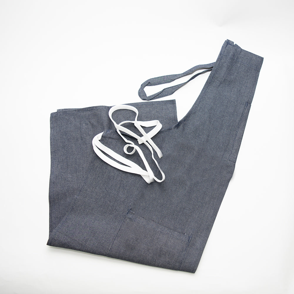 The Calderon Textiles Apron, Denim, is shown laid flat on a white background. This dark gray apron with white straps offers superior comfort and features two large front pockets for stylish, practical use.