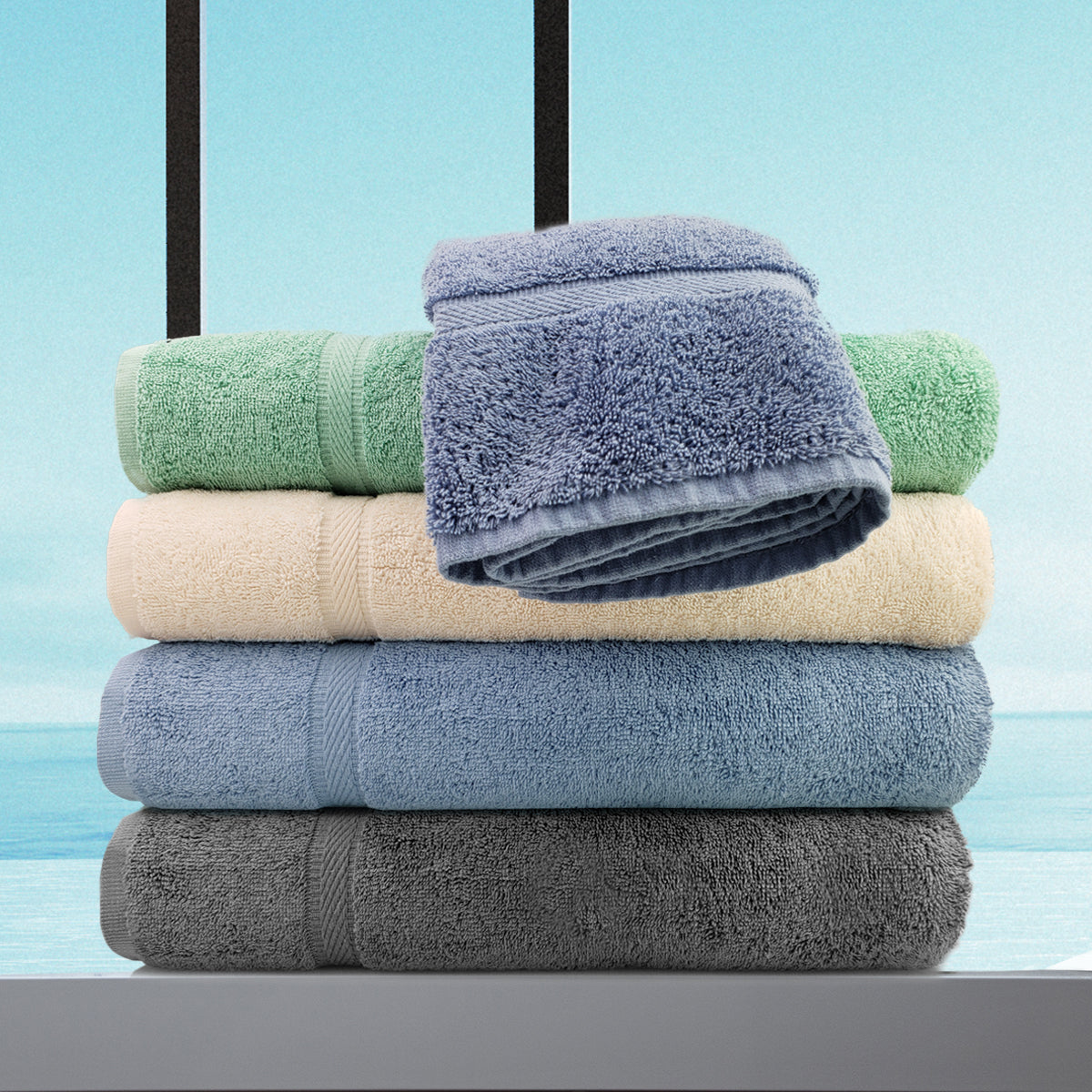 A stack of four folded Ganesh Mills Oxford Imperiale bathmats in green, cream, blue, and gray, with a fifth blue one draped on top. Made from cotton ringspun for absorbency, set against bright sky and water for a serene spa-like vibe.