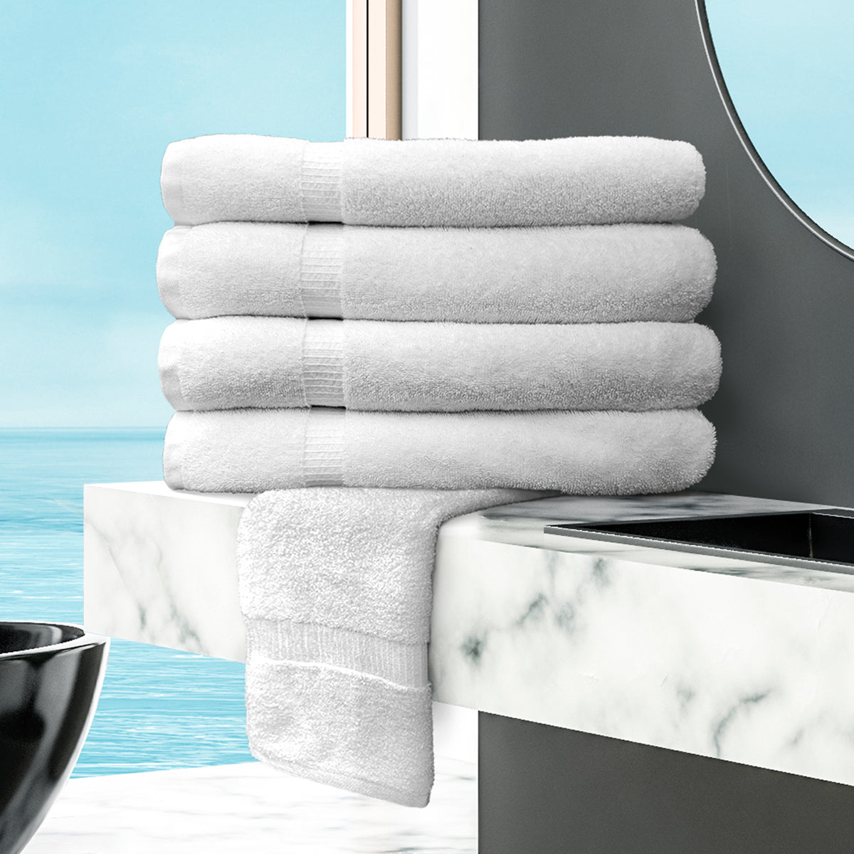 Five Ganesh Mills Oxford Bellezza Bathmats are neatly stacked on a marble bathroom counter, with one draped slightly. A round mirror and ocean view appear in the background.
