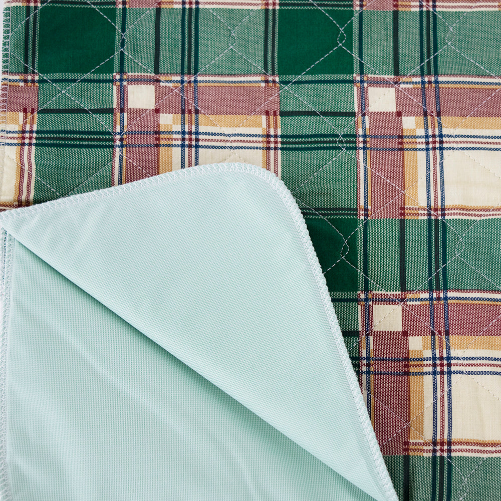 A green and brown plaid fabric with white stitching is partially covered by a corner-folded Calderon Textiles Underpad Vinyl Lining in light blue, size 34 x 36, over the top left side.