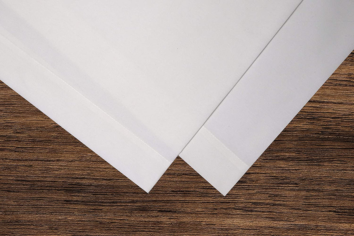 Two white sheets overlap on wood, evoking the crisp, clean look of KSE’s Pillowcase – Premium T130 (Standard) bedding, with a contrast between the smooth “fabric” effect and textured grain.