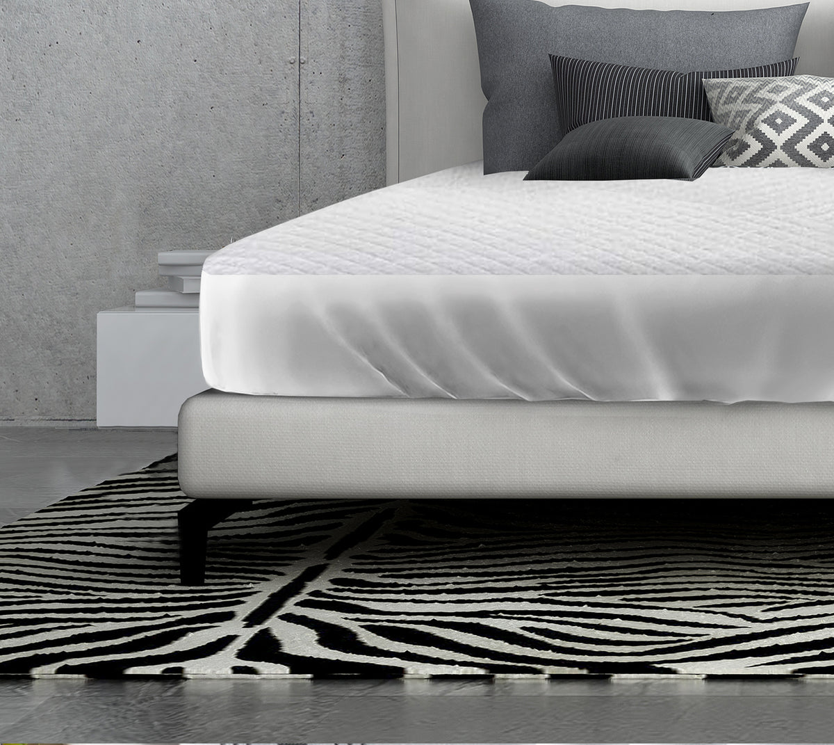 A modern bed with a white mattress protected by the Ganesh Mills Oxford Quilted Bed Pads Fitted, gray and patterned pillows, and a black-and-white geometric rug in a minimalist room with a concrete wall and stacked books on a white nightstand.