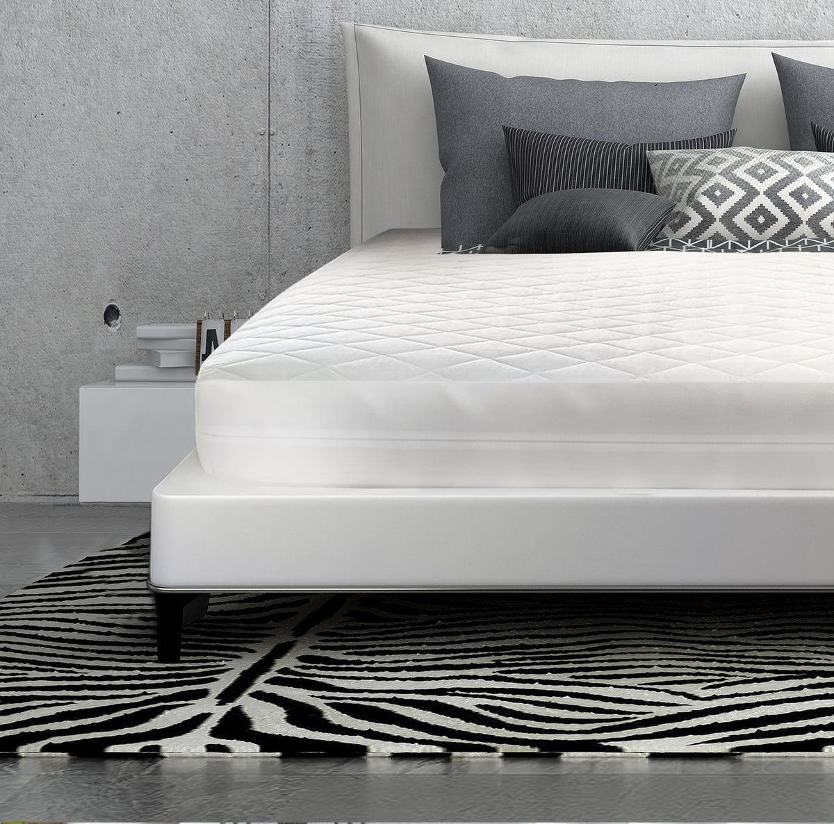 A modern bed with a white mattress topped by the Oxford Three Layer Quilted Bed Pad Waterproof Fitted from Ganesh Mills, gray and patterned pillows, and a geometric black-and-white rug, set against a concrete wall with a white nightstand on the left.