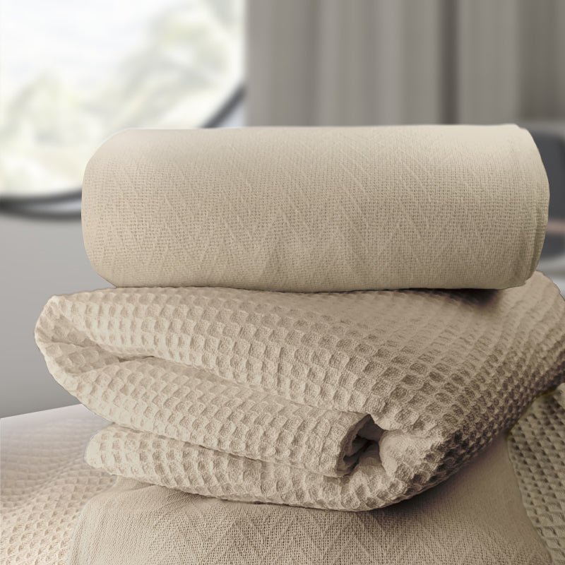 A neatly folded stack of Ganesh Mills Oxford Herringbone Blankets in white/beige rests on a surface, illuminated by soft natural light through a window with neutral curtains in the background.