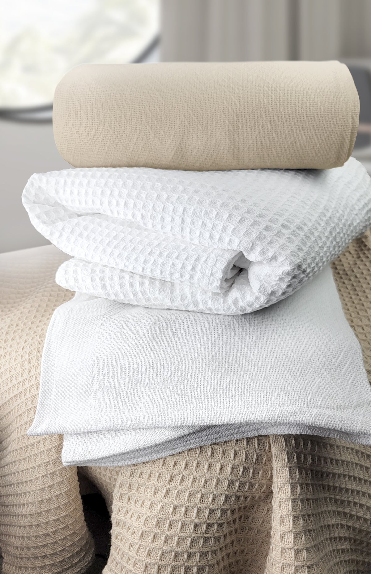 A stack of white and beige textured towels rests on a Ganesh Mills Oxford Jaipur Big Honeycomb Blanket in white/beige, featuring a waffle pattern, set in a bright room with blurred windows in the background.