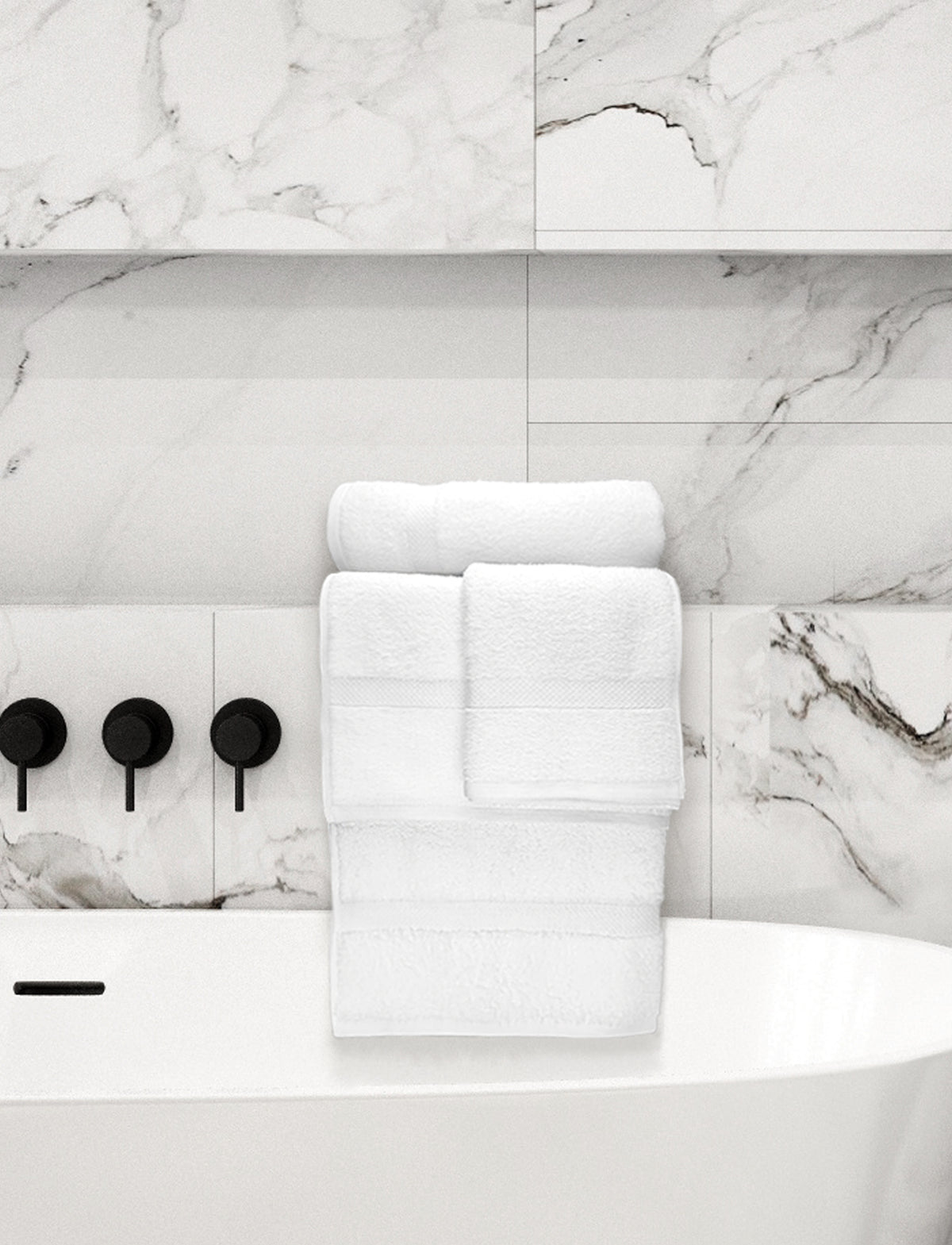 A stack of Ganesh Mills Washcloth - White Oxford Nuvola Towels is neatly arranged on the edge of a white bathtub, set against white marble tiles with gray veining and matte black faucet handles.