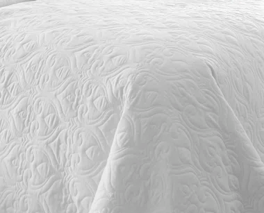 A close-up of the KSE Ribcord Bedspread, featuring a white quilted design with an embossed, ornate swirling pattern in fire-retardant polyester—perfect for bulk hospitality use.