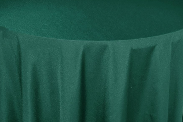 A close-up of a round table draped in a dark green, floor-length Ganesh Mills Tablecloth - Oxford Blend Satin Band Table Linen, showcasing soft vertical folds that evoke the sophisticated look seen at formal events.