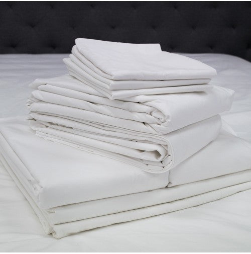 A stack of Intralin Pillowcase - T-130 Econolin sheets, import, made from a cotton-polyester blend, is neatly folded on a white bed with a dark tufted headboard in the background.