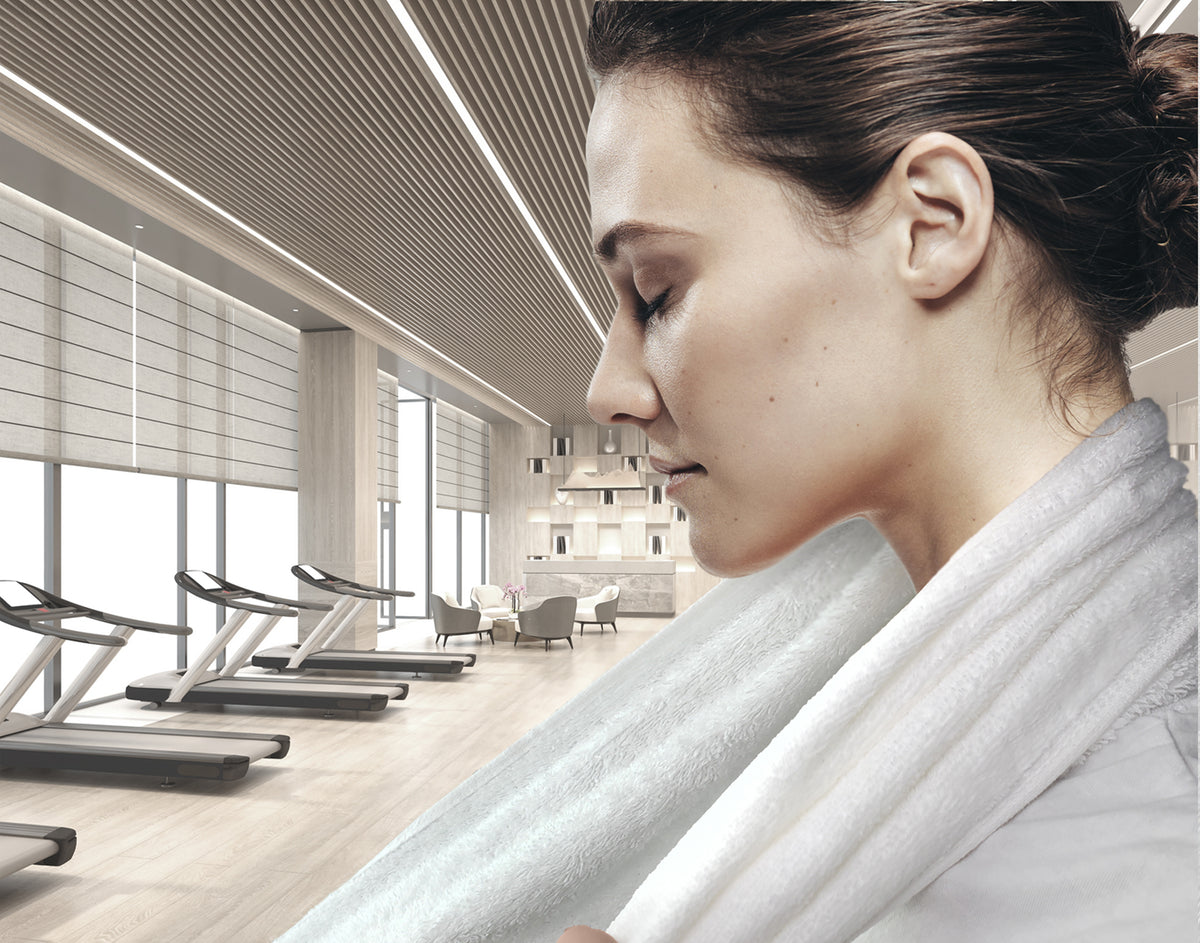 A woman stands in profile at a modern gym with large windows and light wood decor, wearing a Ganesh Mills Spa Velour Hand Towel - White around her neck.