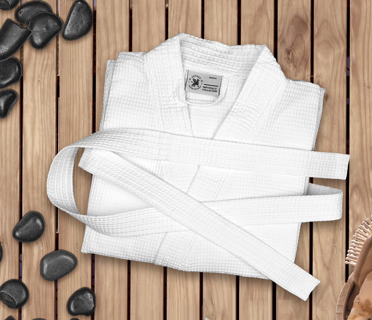 A neatly folded white martial arts uniform with a belt rests on a wooden surface, flanked by black spa stones and a Ganesh Mills Oxford Honeycomb Waffle Bathrobe in white 100% cotton, along with a wooden bath brush.