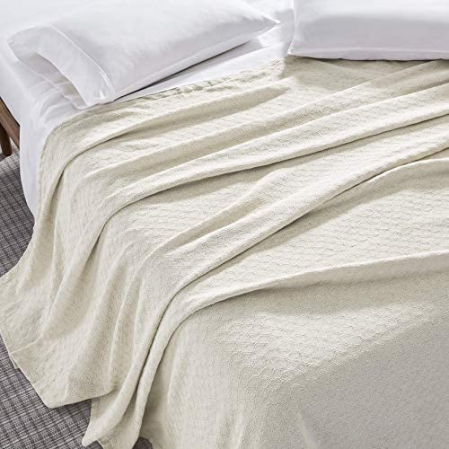 A neatly made bed features white pillows and an Intralin Bath Blanket in light beige, casually draped over the mattress and partially covering a gray carpeted floor.