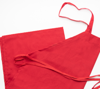 A Prisma Bib Apron by Calderon Textiles in bright red polyester is laid flat with a matching red napkin on a white surface, showing the apron’s neck strap and waist ties for added comfort.