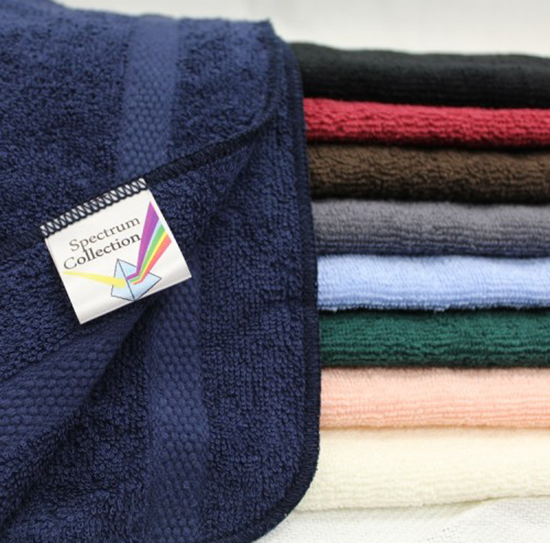 A stack of Intralin Spa Hand Towel - Spectrum Towels in navy blue, black, brown, gray, light blue, pink, peach, and cream is neatly folded. The top soft towel displays the Spectrum Collection label.