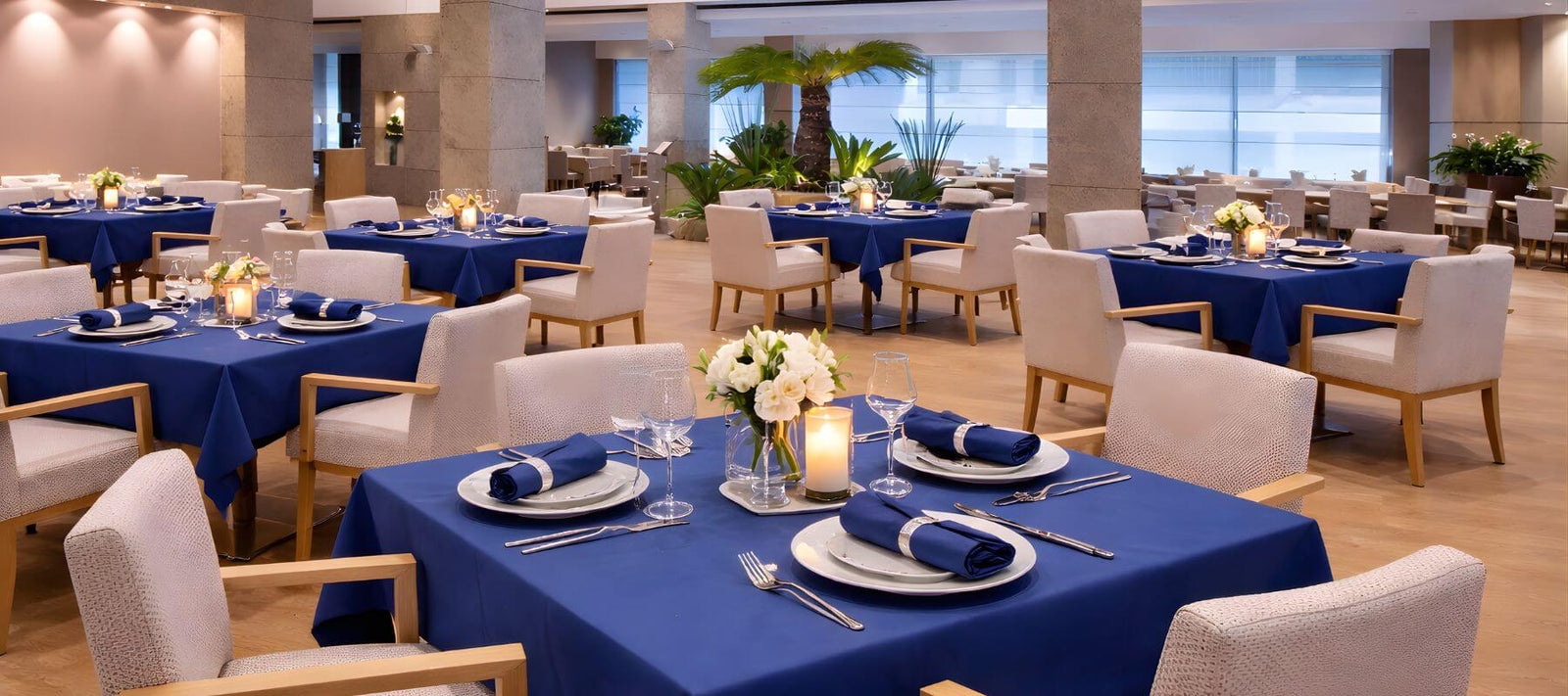 A modern restaurant with blue tablecloths, white cushioned chairs, flower centerpieces, and candles on each table. Large windows and plants create a bright, elegant atmosphere.