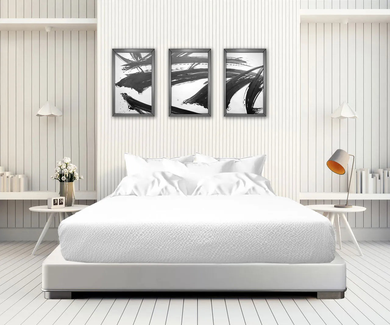 A minimalist bedroom features a white bed with the Ganesh Mills Oxford Bed Topper - Crinkle Weave, side tables, modern lamps, and three abstract black-and-white paintings above. White walls, shelves with books, and a vase of flowers complete the look.