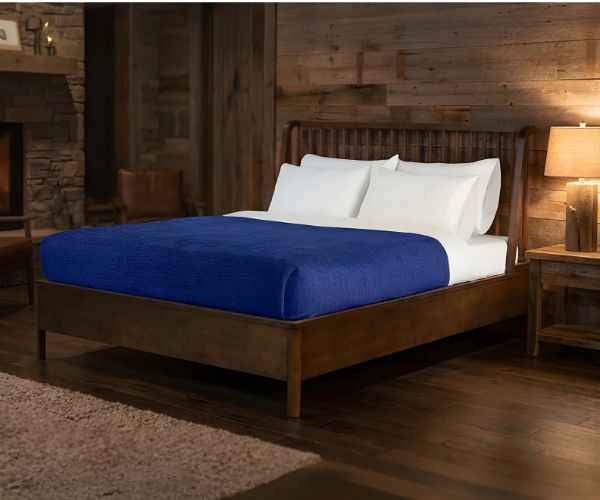 A wooden bed with white pillows and a cozy WestPoint Home Vellux Renew Chenille Blanket sits in a rustic bedroom featuring wood-paneled walls, a nightstand with a lamp, and a stone fireplace in the background.