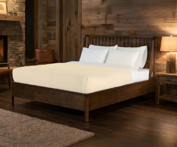 A cozy bedroom with a wooden bed frame, white bedding, and the WestPoint Home Vellux Renew Chenille Blanket. Wood-paneled walls, a lamp-topped nightstand, soft rug, and stone fireplace complete the warm, rustic atmosphere.