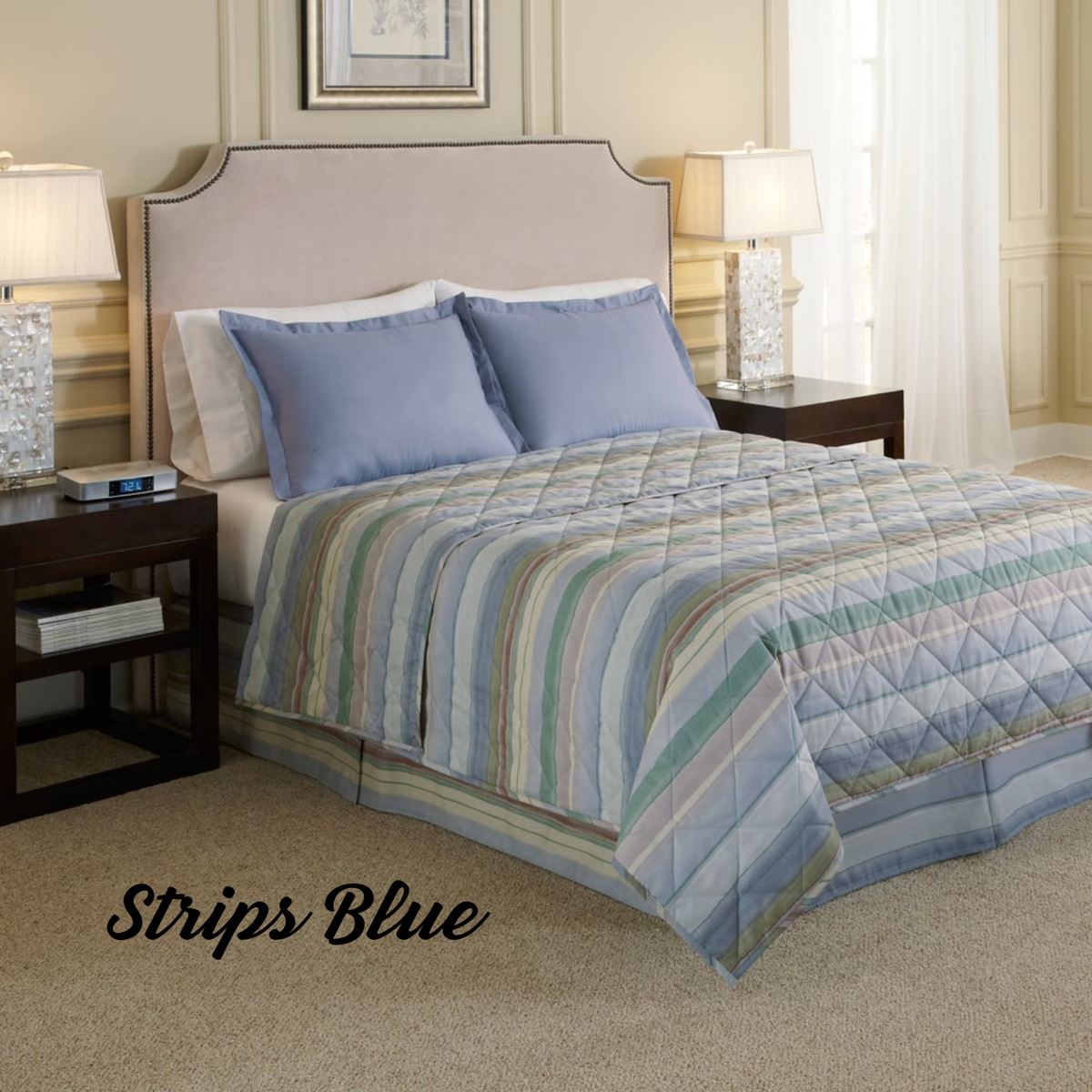 A neatly made bed with a light beige headboard, blue pillows, and a WestPoint Hospitality "Pattern" comforter by WestPoint Home creates a luxury hotel look. Nightstands with lamps and a clock sit on either side. “Strips Blue” appears in the lower left corner.