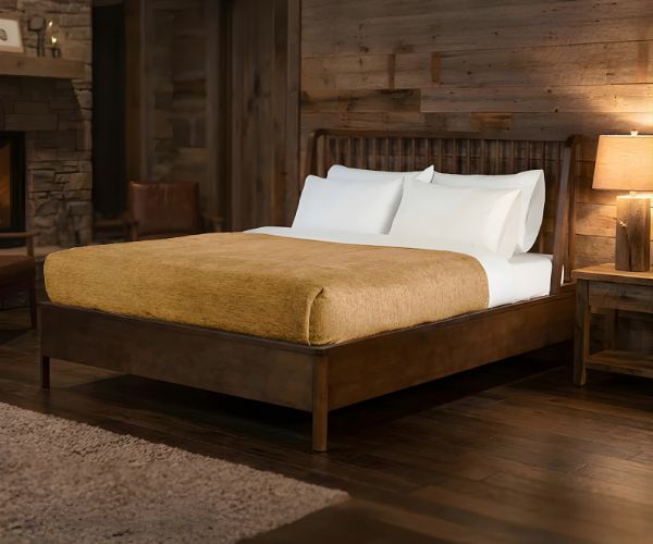 A cozy bedroom with a wooden bed frame, white pillows, a beige WestPoint Home Vellux Renew Chenille Blanket, a wooden nightstand with lamp, and wood-paneled walls creates a warm, rustic atmosphere.