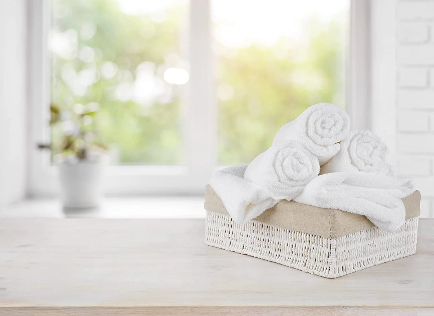 A wicker basket with neatly rolled KSE Bath Towel - Basic Economy 10 Single Towels sits on a wooden surface, with a blurred bright window and a potted plant in the background.