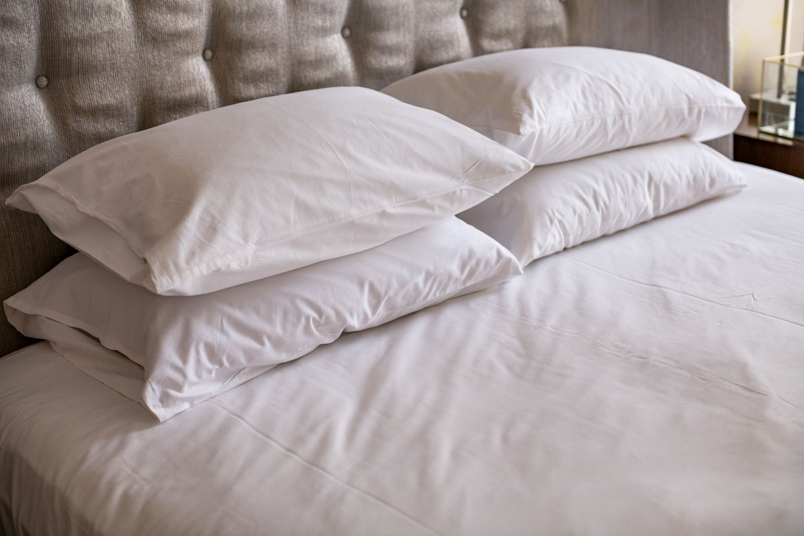 Close-up of a neatly made bed featuring four stacked white pillows and crisp Thomaston Mills Healthcare sheets - T-180 Impressions Print, set against a tufted, upholstered headboard.