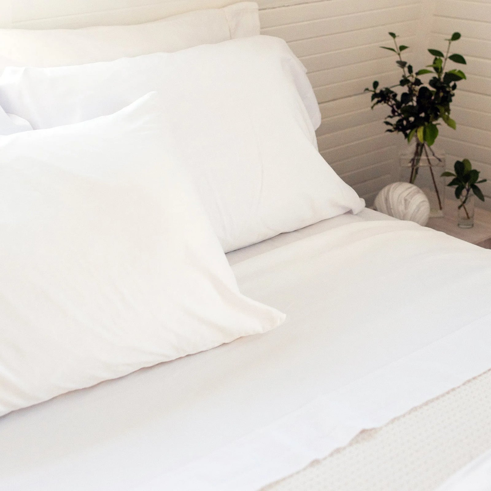A neatly made bed with white sheets and a Berry Compliant Pillowcase USA Bedding sits beside a table with a vase of green leafy plants and a decorative object, all set against a white paneled wall.