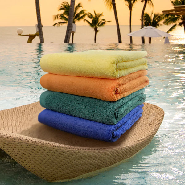 A stack of four Oxford B2700 Premium Pool Towels from Ganesh Mills—yellow, orange, teal, and blue—rests on a woven lounge chair by the pool with palm trees and a sunset ocean view in the background.