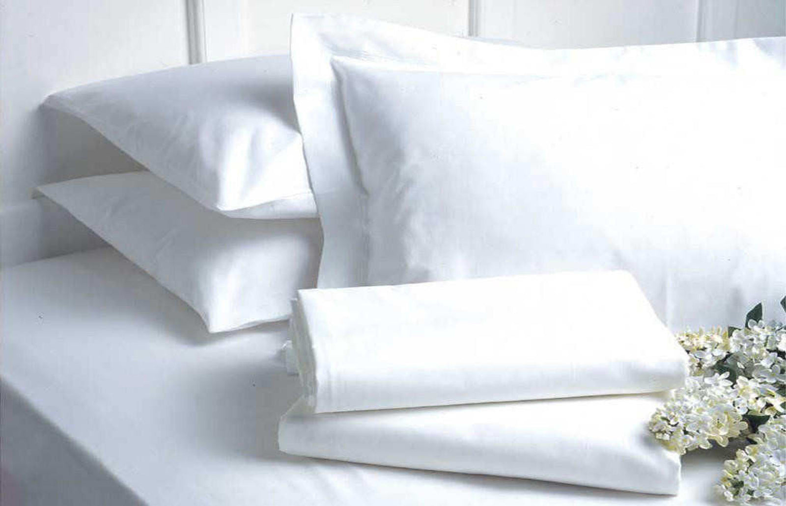 A neatly made bed with crisp Ganesh Mills Pillow Case 4 Hem – Oxford Micro Superblend Bed Linen and smooth white sheets is adorned with folded linens and small white flowers, creating a clean, fresh look.